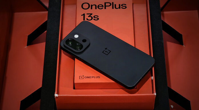Matte black OnePlus 13s placed diagonally on top of its signature red retail box, highlighting the bold rear camera design and premium build.