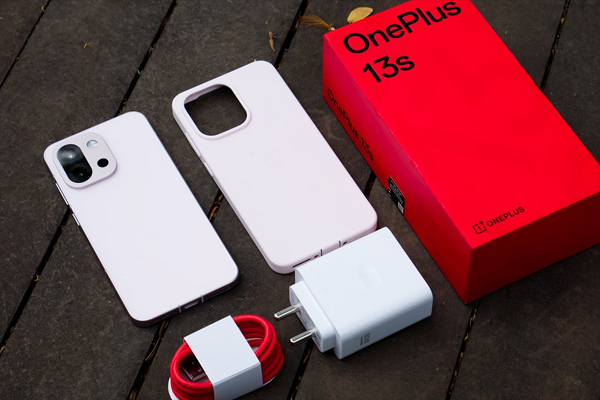 OnePlus 13s unboxing showing the smartphone, transparent TPU case, 100W SUPERVOOC charger, USB Type-C cable, and red retail box laid out on a wooden surface.